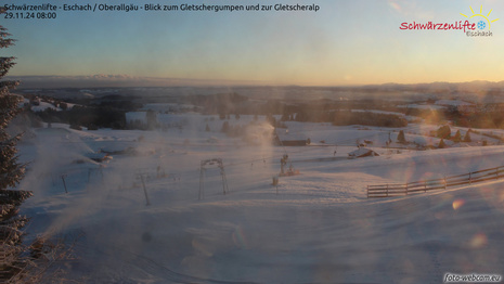 Webcamfoto zeigt schneebedeckte Landschaft im Allgäu bei Sonnenaufgang sowie laufende Beschneiungsanlagen im Pistenbereich. (Quelle www.foto-webcam.eu)