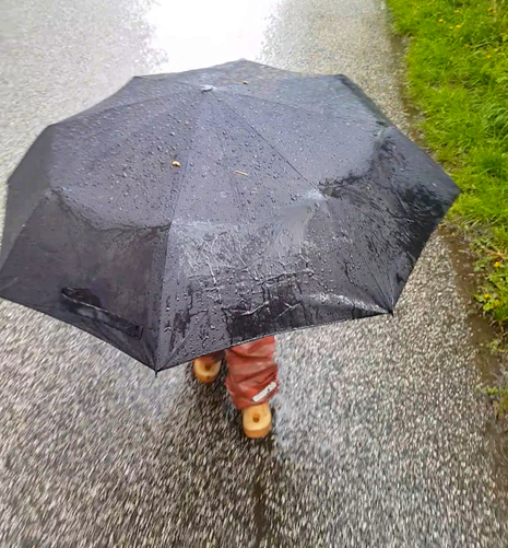 Das Foto zeigt einen regnerischen Tag und ein Kind in Gummistiefeln und Regenhose unter einem großen schwarzen Regenschirm