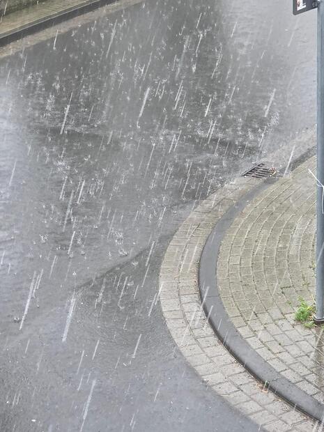 Foto zeigt große Regentropfen, die auf eine Straße und einen Gehweg fallen. (Quelle DWD - Deutscher Wetterdienst)