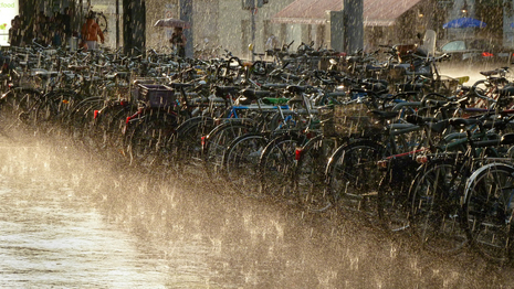 Bild zeigt abgestellte Fahrräder im sommerlichen Regen in der Abendsonne (Quelle Photones, CC BY-SA 3.0 <https://creativecommons.org/licenses/by-sa/3.0>, via Wikimedia Commons)